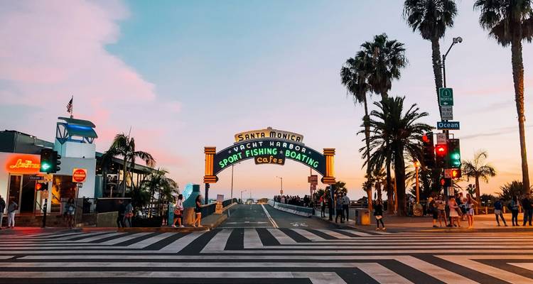 The iconic Santa Monica Pier entrance sign lit at dusk as pedestrians cross a striped intersection.