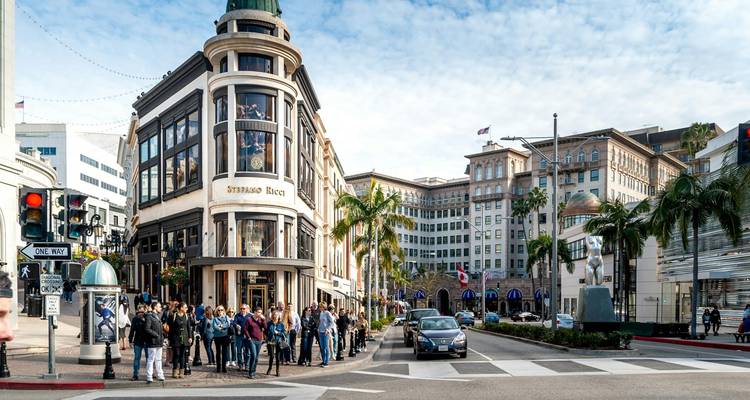 Busy Rodeo Drive intersection with stylish buildings, palm trees and a crowd of shoppers waiting to cross.