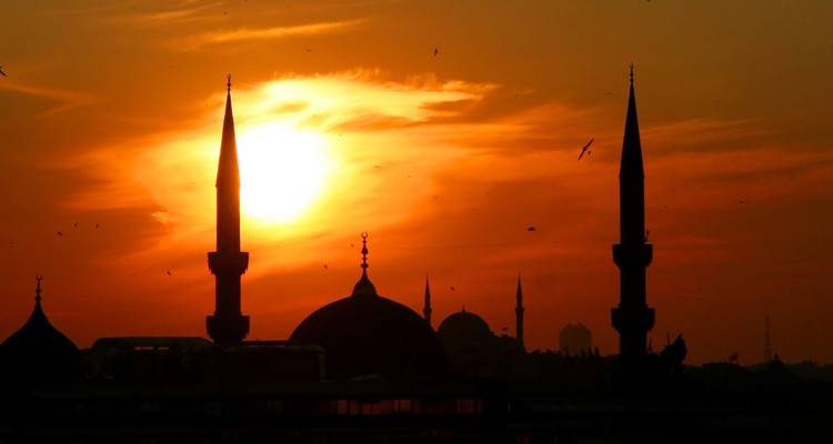 Silhouet van koepels en slanke minaretten tegen een oranje zonsonderganghemel met vliegende vogels.