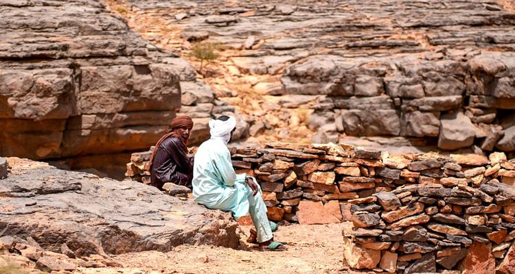 Two Tuareg men sit on stone walls in a rugged, rocky desert landscape chatting.
