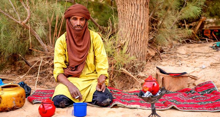 A Tuareg man in yellow robe prepares tea on a patterned rug beneath desert trees.
