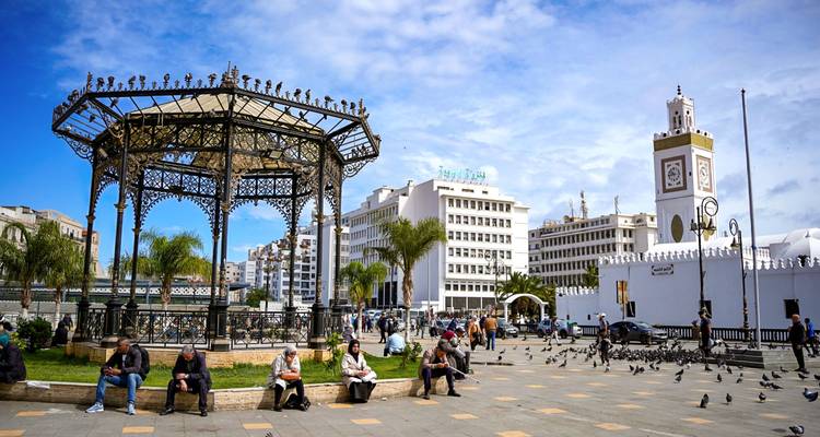 City square with ornate iron gazebo, people relaxing on benches, pigeons, and a white clock-tower mosque nearby.