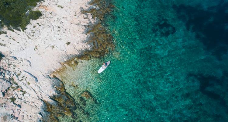 Top-down aerial view of a paddleboarder gliding over crystal-clear turquoise water along a rocky Croatian shoreline.