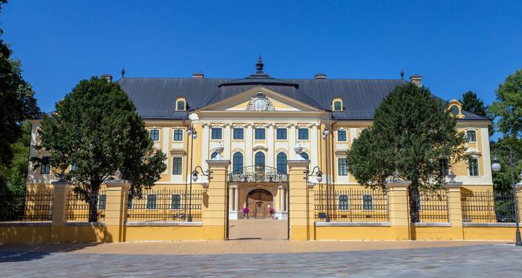 Elegant yellow baroque palace fronted by tall trimmed trees and an iron fence against a clear blue sky.