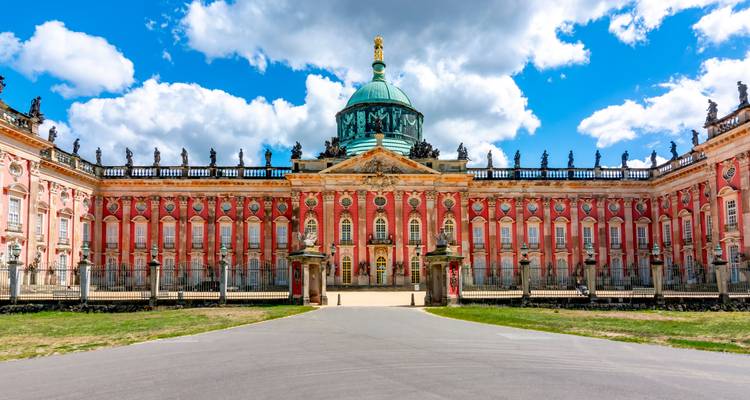 Ornate red-and-gold façade of Potsdam’s New Palace crowned by a green copper dome beneath dramatic clouds.