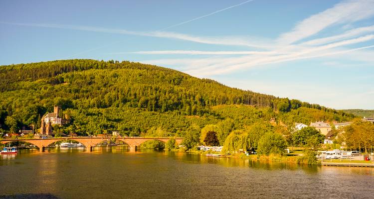 Tranquil river scene with a red-brick arched bridge, forested hills and scattered riverside houses in soft light.