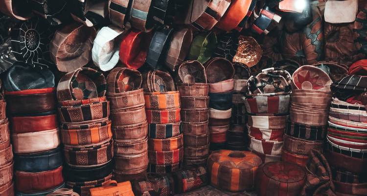 Stacks of colorful leather poufs and bags fill a dim Moroccan souk stall.