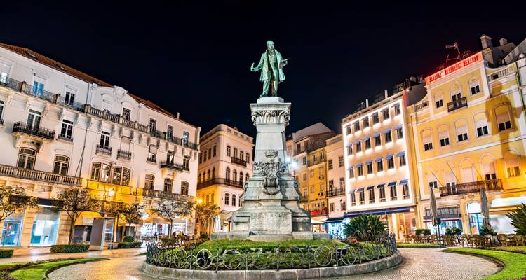 Illuminated statue in a lively European square at night surrounded by historic buildings.