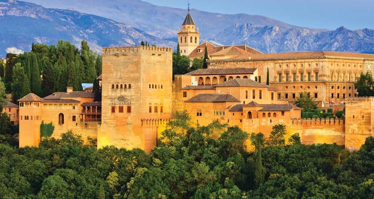 Sun-lit fortress complex of the Alhambra perched above dense trees with mountains in the background.