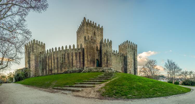 Medieval Guimaraes Castle with crenellated stone walls sits atop a grassy mound under a clear sky.
