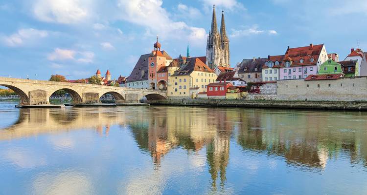 Stone bridge, pastel riverfront houses and twin spires of Regensburg reflected in calm Danube waters.