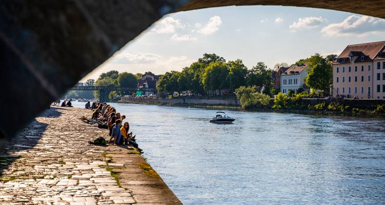 Locals relax on stone embankment of a wide river viewed through an arching bridge.