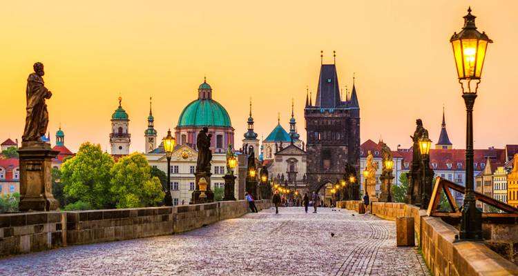 Historic Charles Bridge in Prague at sunrise with statues and glowing lanterns leading to the Old Town skyline.