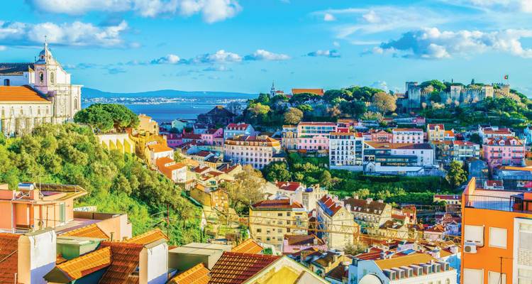 Colorful rooftops of Lisbon’s Alfama district cascading toward the Tagus River with castle and church on the horizon.