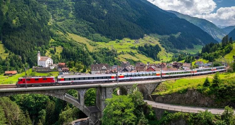 Sleek red train crosses a high stone viaduct in a verdant Swiss alpine valley dotted with villages.
