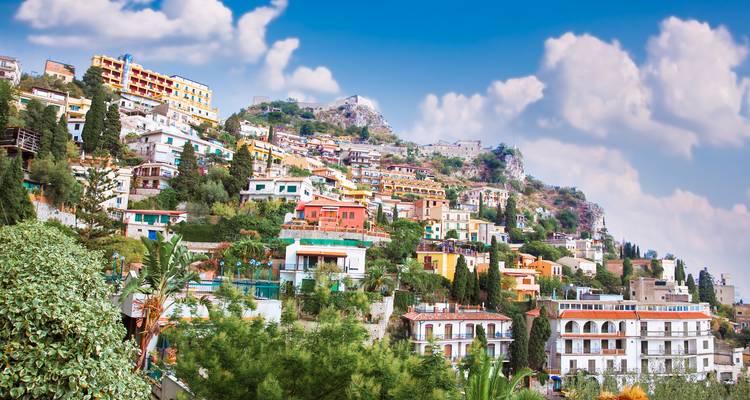 Colorful houses cascade down a lush hillside of a Mediterranean coastal town under a bright blue sky with fluffy clouds.