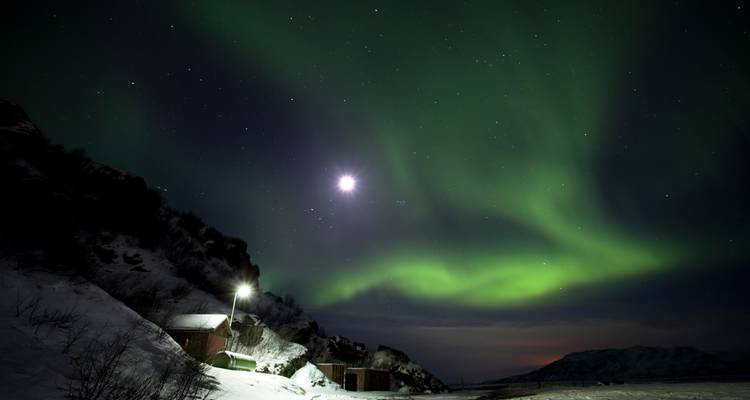 Une aurore verte tourbillonne dans un ciel d'hiver étoilé au-dessus d'une cabane enneigée et de montagnes illuminées par la lumière lunaire.