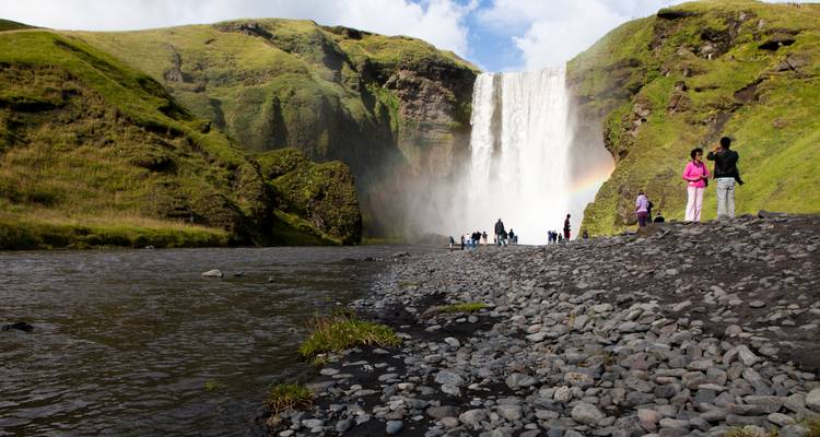 Les visiteurs se rassemblent à la base de Skógafoss où la brume et la lumière du soleil créent un léger arc-en-ciel au-dessus de la rive.