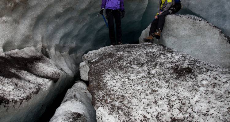 Gros plan de deux trekkeurs en équilibre sur la glace granuleuse à l'intérieur d'une crevasse de glacier montrant des parois ondulées de couleur bleue.