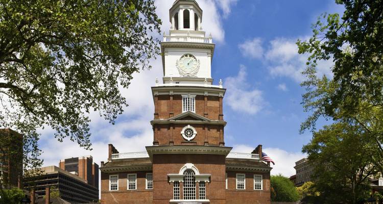 Brick façade of Independence Hall with white clock tower framed by green trees against blue sky.