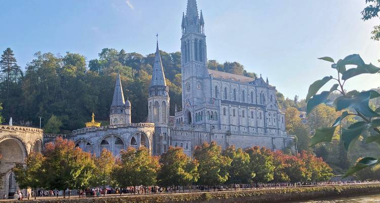 Santuario de Lourdes elevándose sobre la orilla del río bordeada de árboles con multitudes de peregrinos abajo.