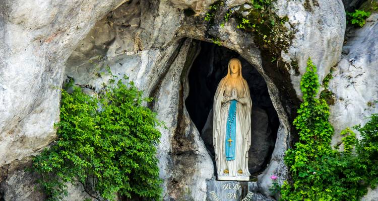 Estatua de la Virgen María en la gruta de Lourdes rodeada de exuberante vegetación.