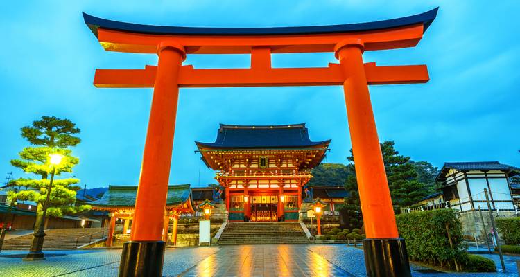 Iconic vermilion torii gate standing before an illuminated Shinto shrine at dusk