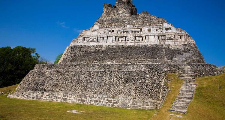 Grand temple Maya en pierre avec des frises complexes au sommet d'une butte herbeuse sous un ciel bleu sans nuages.