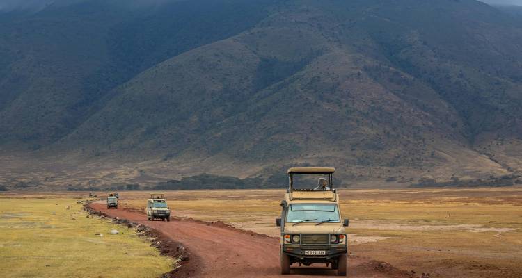 Series of safari vehicles driving along a red dirt track across open plains toward a mountain