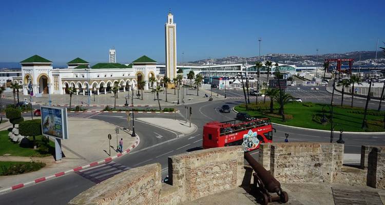 Breiter Küstenplatz mit gewölbtem Terminalgebäude, rotem Sightseeing-Bus und entfernter Bucht unter strahlend blauem Himmel.