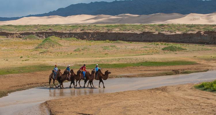 Small camel caravan crossing a shallow river with towering sand dunes and mountains beyond