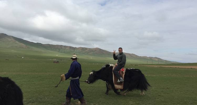 Tourist riding a yak led by a Mongolian herder across open grassland under cloudy skies