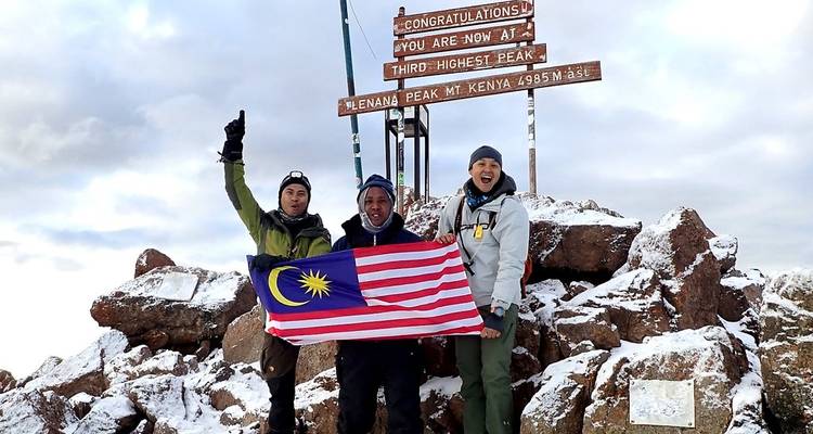 Three climbers celebrating on snowy Point Lenana summit holding a Malaysian flag beside a wooden congratulatory sign