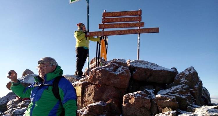 Climbers standing on a rocky, snow-dusted summit platform with a sign marking Point Lenana under a clear blue sky