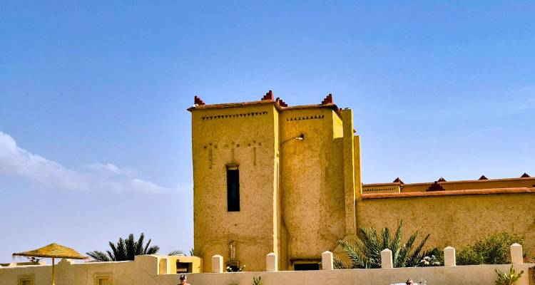 Edificio simple de color amarillo ocre con estilo de kasbah bajo un cielo azul despejado con palmeras en primer plano.