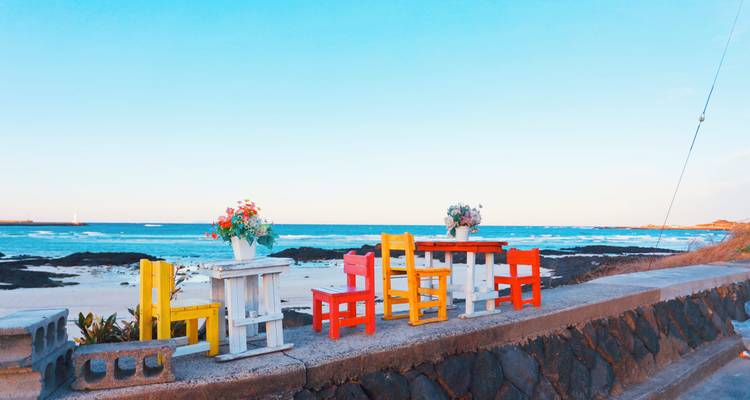 Leuchtend bemalte Stühle und Tische blicken auf einen ruhigen türkisfarbenen Strand auf Jeju.