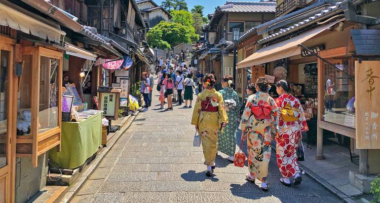 Mujeres vestidas con kimono caminando por una calle comercial tradicional en Kioto llena de tiendas de madera y turistas.