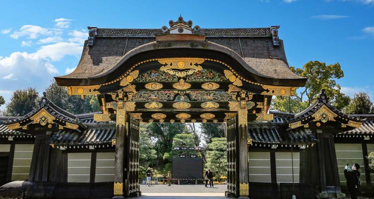 Puerta de entrada del Castillo de Nijo, decorada de forma lujosa y reluciente bajo un cielo azul con algunos visitantes cerca.