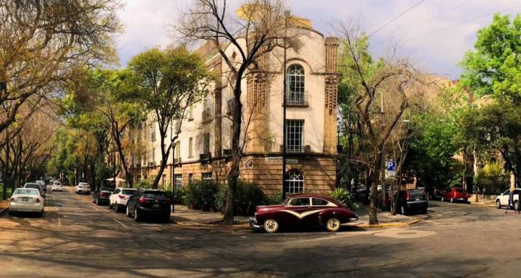 Ruhige, von Bäumen gesäumte Nachbarschaftsstraße mit historischem Apartmenthaus und vintage bordeauxrotem Auto am Straßenrand geparkt.
