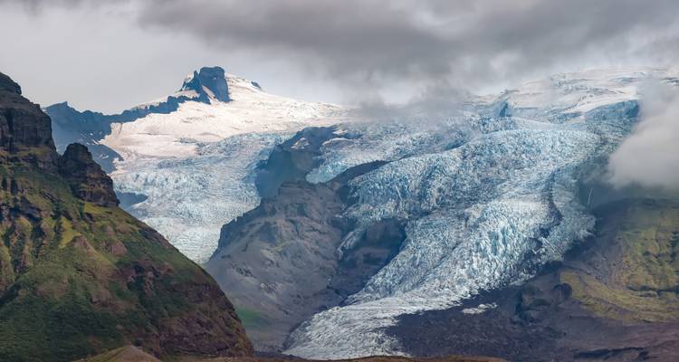 Enorme gletsjer die stroomt tussen ruwe groene en vulkanische bergen onder wolken