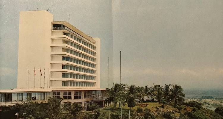 Mid-century high-rise hotel overlooking tropical gardens and coastline under an overcast sky.
