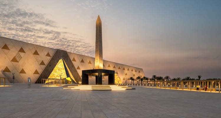 Modern museum facade with illuminated obelisk and geometric wall panels at twilight