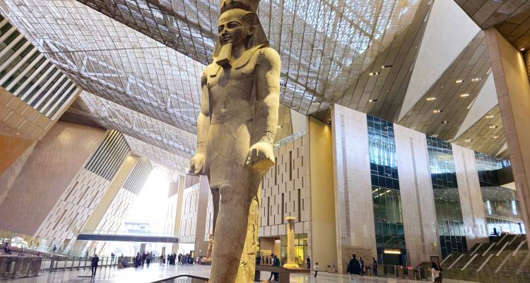 Colossal ancient statue inside a vast glass-roofed museum atrium with visitors below