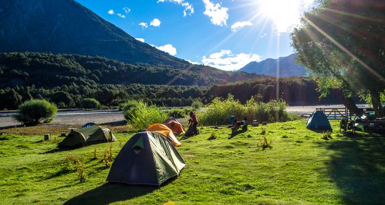 Grassy riverside campsite dotted with colorful tents and sunbeams streaming over mountains