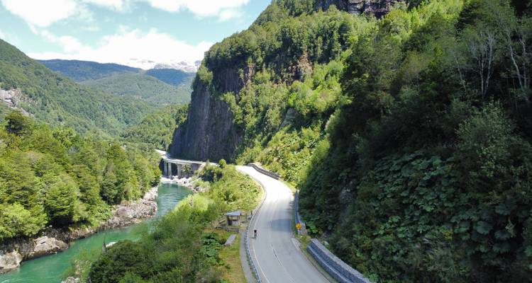 Curving mountain roadway running beside a jade-green river in a steep forested gorge with snowy peaks in the distance.
