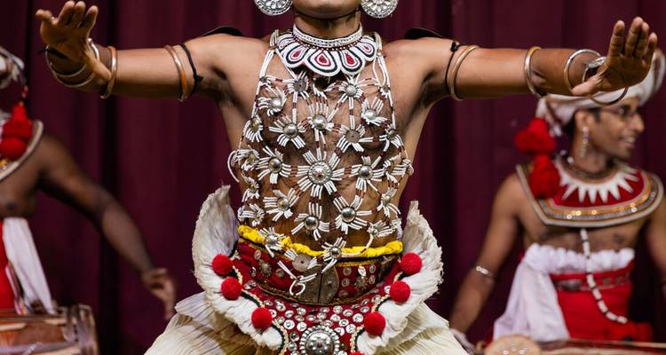 Traditional Kandyan dancer in ornate silver chest piece performing with drummers on stage.