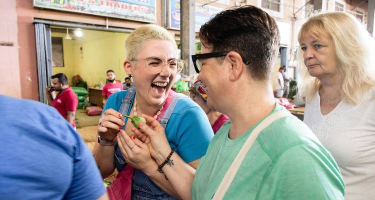 Tourists laugh and sample produce in a bustling local market aisle.