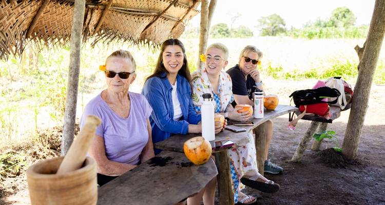 Four travellers sit on a rustic wooden bench under a thatched shelter, sipping fresh coconuts in a sunny rural setting.