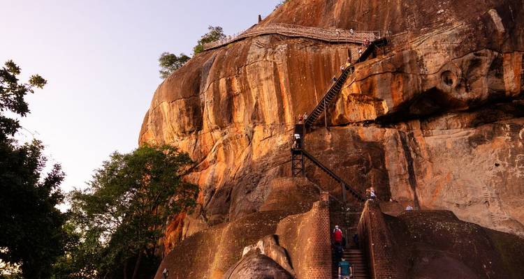 Visitors ascend the narrow stairs on the face of the massive Sigiriya Rock fortress glowing in warm evening light.