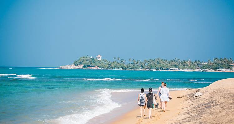 Three women stroll along a tranquil tropical beach beside turquoise waves and a palm-fringed headland.